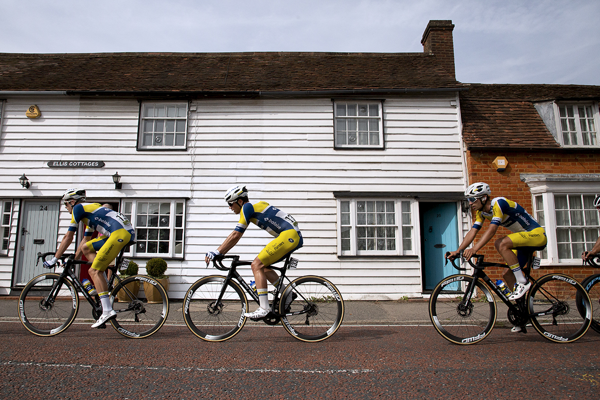 Tour of Britain 2023 - A group of riders pass a row of white wooden cottages with red, white and blue doors