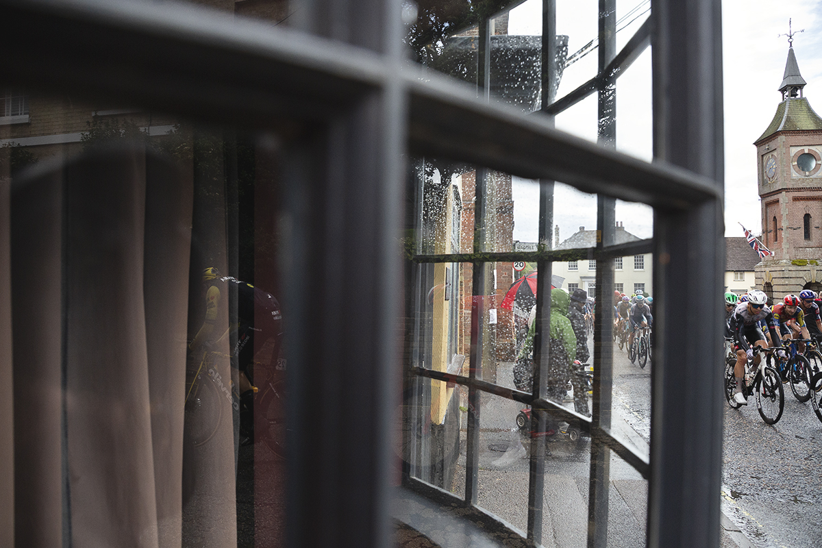Tour of Britain 2025 - Riders are seen through a bay window passing through the market place of Bildeston