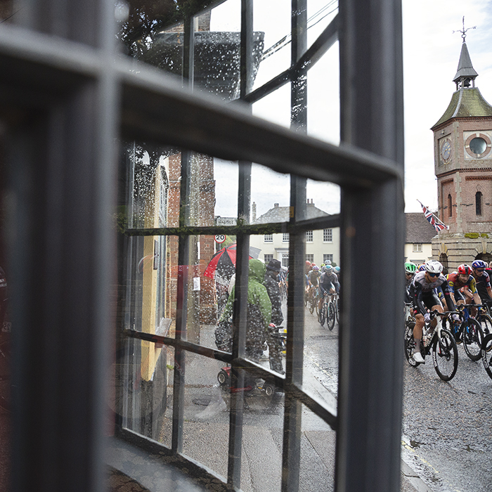 Tour of Britain 2025 - Riders are seen through a bay window passing through the market place of Bildeston