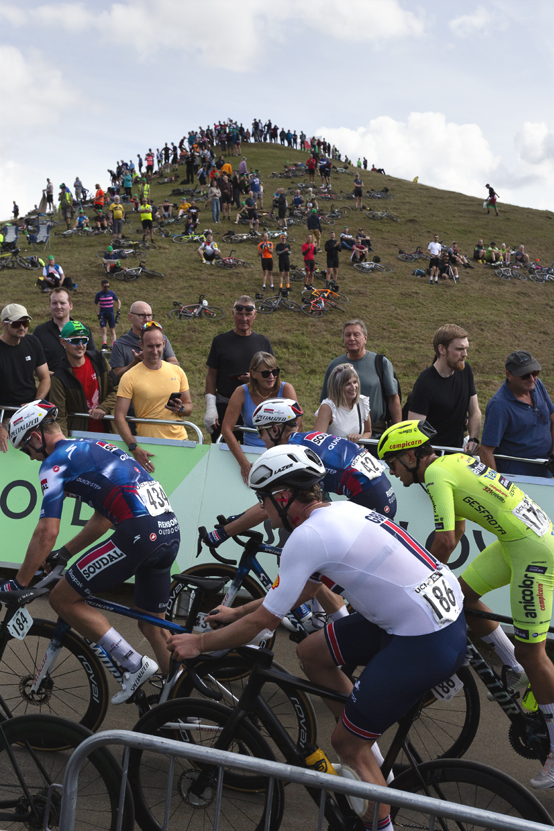 Tour of Britain 2025 - Fans bang the hoardings while in the background the hummock of a hill in lined with fans at Burton Dassett