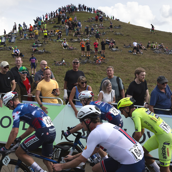 Tour of Britain 2025 - Fans bang the hoardings while in the background the hummock of a hill in lined with fans at Burton Dassett