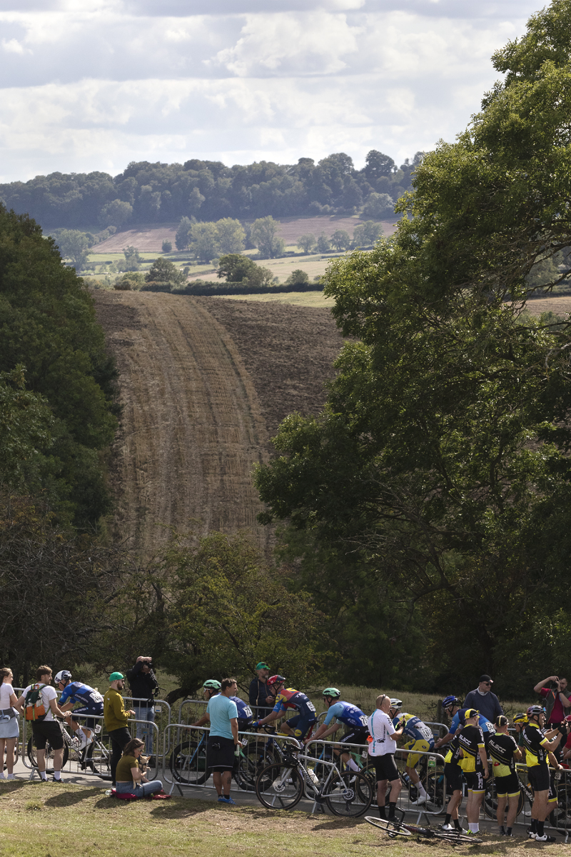 Tour of Britain 2025 - Riders pass groups of fans at the roadside in Burton Dassett with countryside behind them
