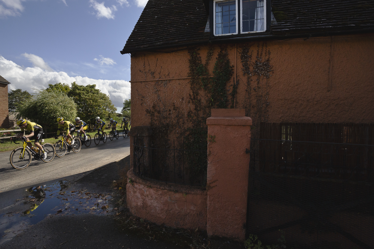 Tour of Britain 2025 - Riders are reflected in a puddle as they pass a terracotta painted house in Cople