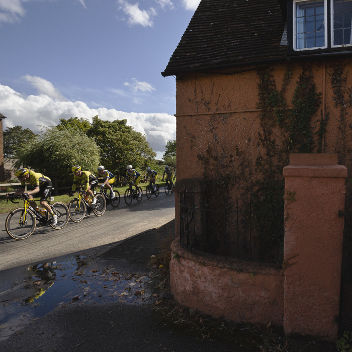 Tour of Britain 2025 - Riders are reflected in a puddle as they pass a terracotta painted house in Cople