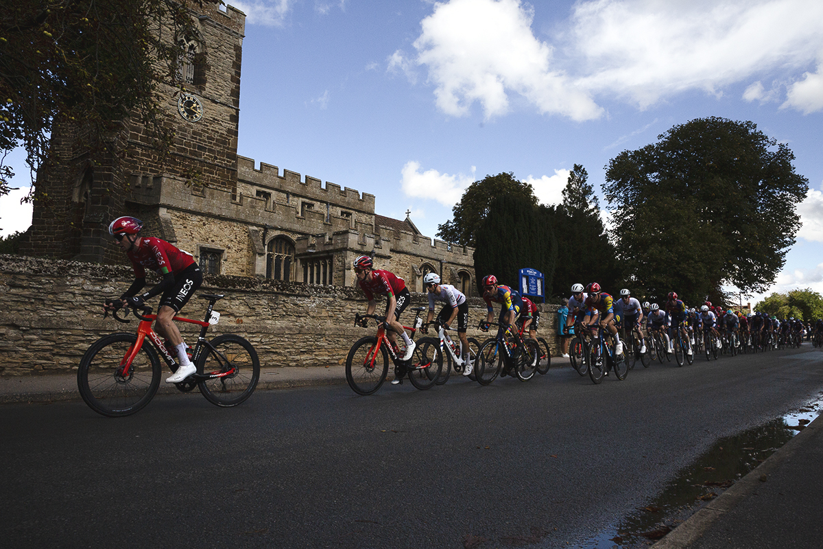 Tour of Britain 2025 - A line of riders pass in front of the church in Cople
