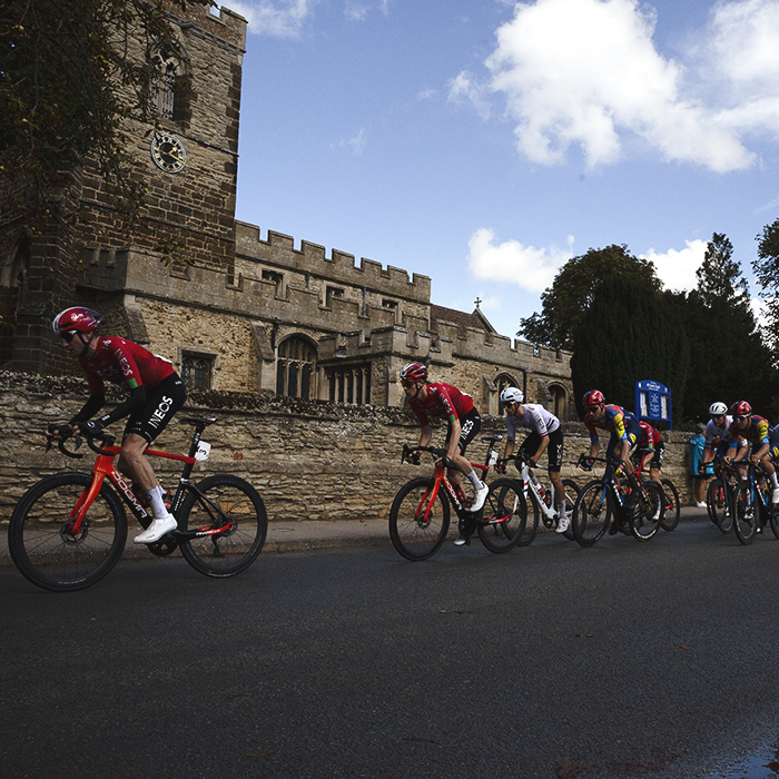 Tour of Britain 2025 - A line of riders pass in front of the church in Cople
