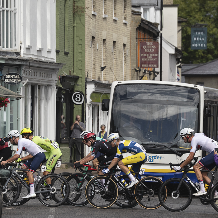 Tour of Britain 2025 - Riders cross a junction in Saxmundham while a local bus service is held up by the race