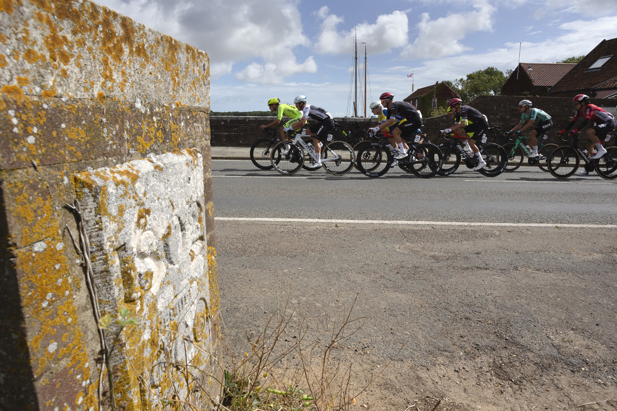 Tour of Britain 2025 - A group of riders make their way onto the bridge at Snape, a foundation stone with the date of the bridge in the foreground