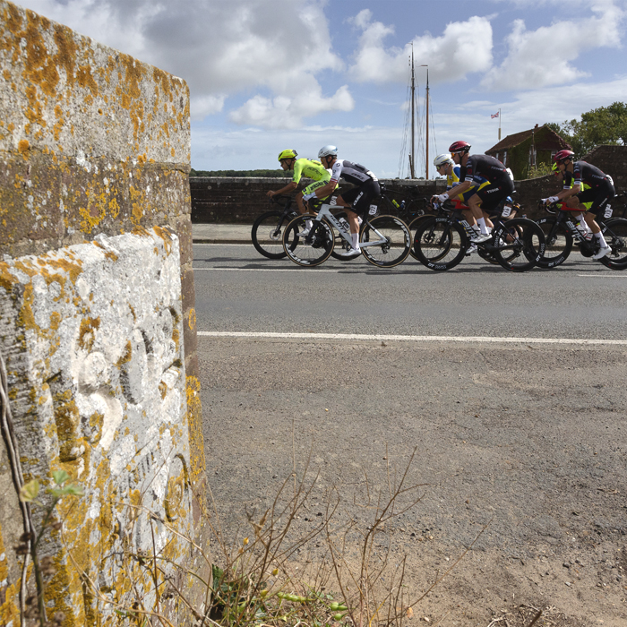 Tour of Britain 2025 - A group of riders make their way onto the bridge at Snape, a foundation stone with the date of the bridge in the foreground