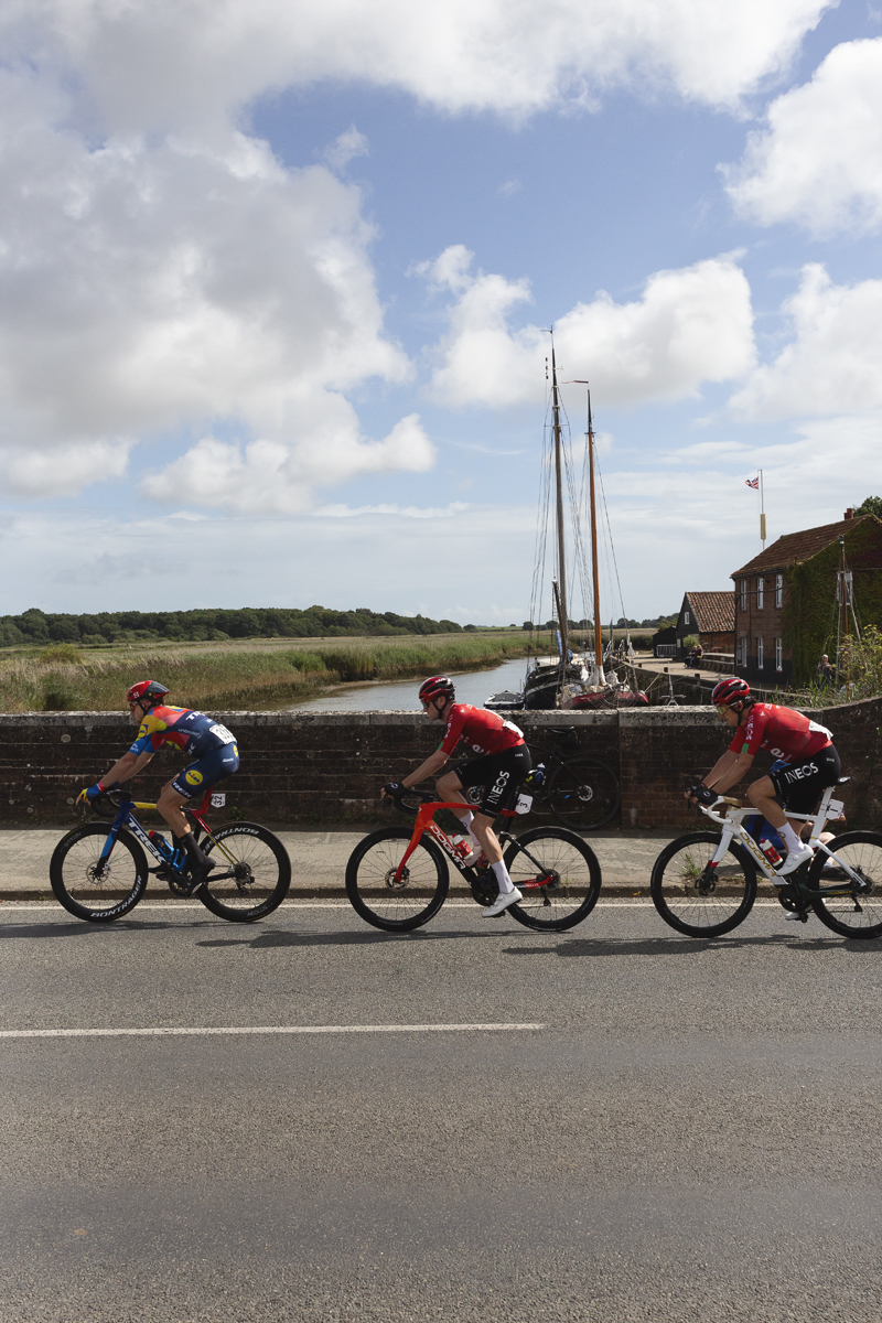 Tour of Britain 2025 - Riders cross the bridge at Snape Maltings with a masted boat in the background