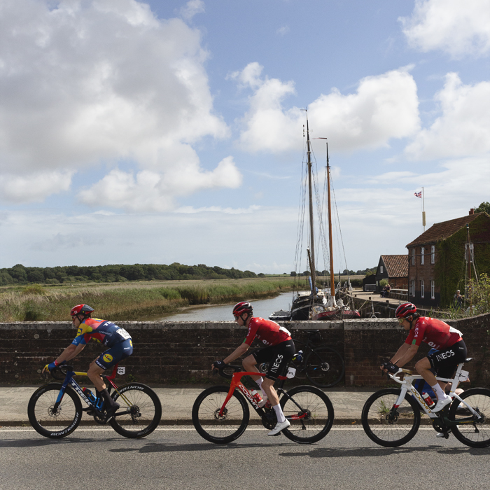 Tour of Britain 2025 - Riders cross the bridge at Snape Maltings with a masted boat in the background