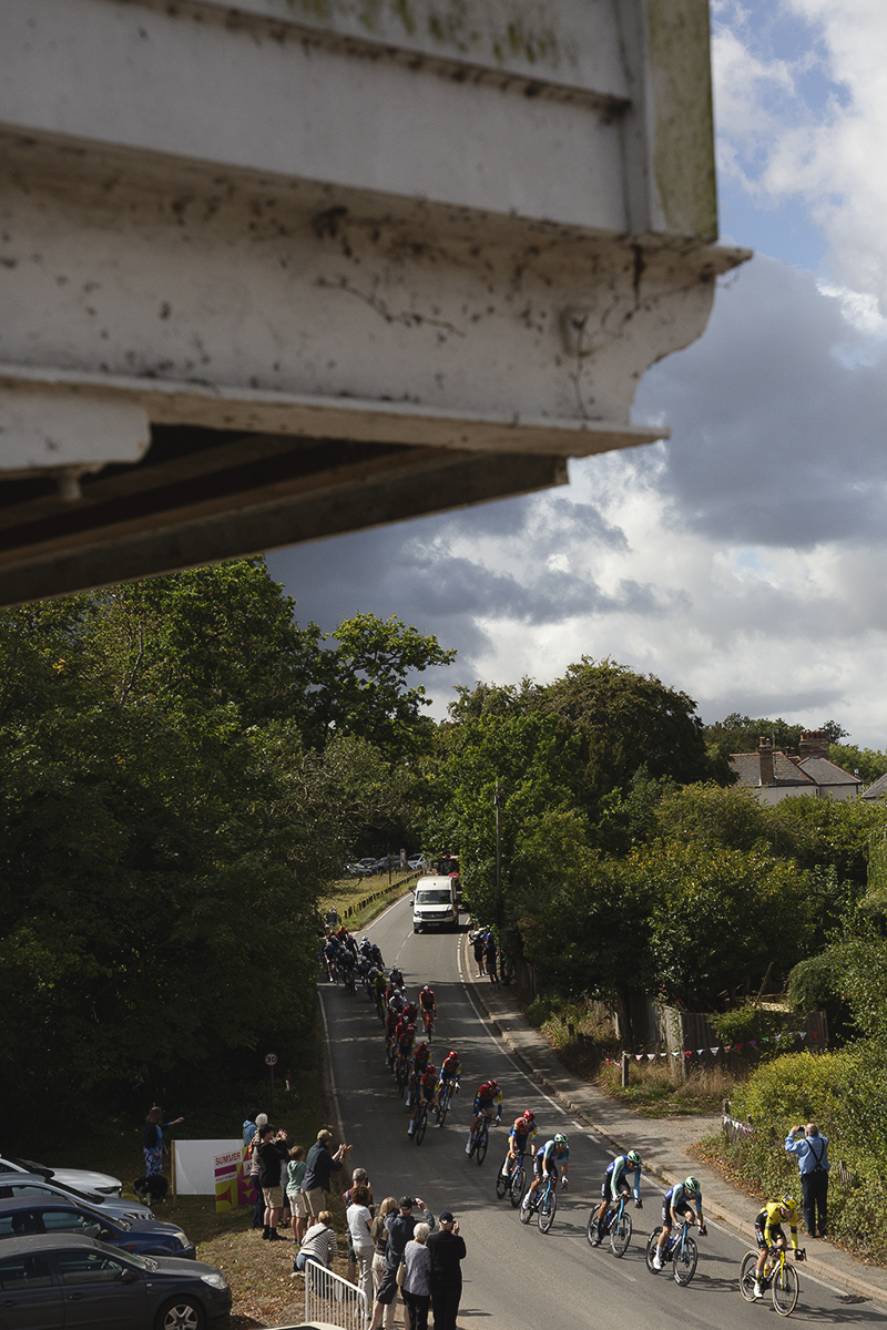 Tour of Britain 2025 - The peloton below the old warehouse buildings at Snape Maltings