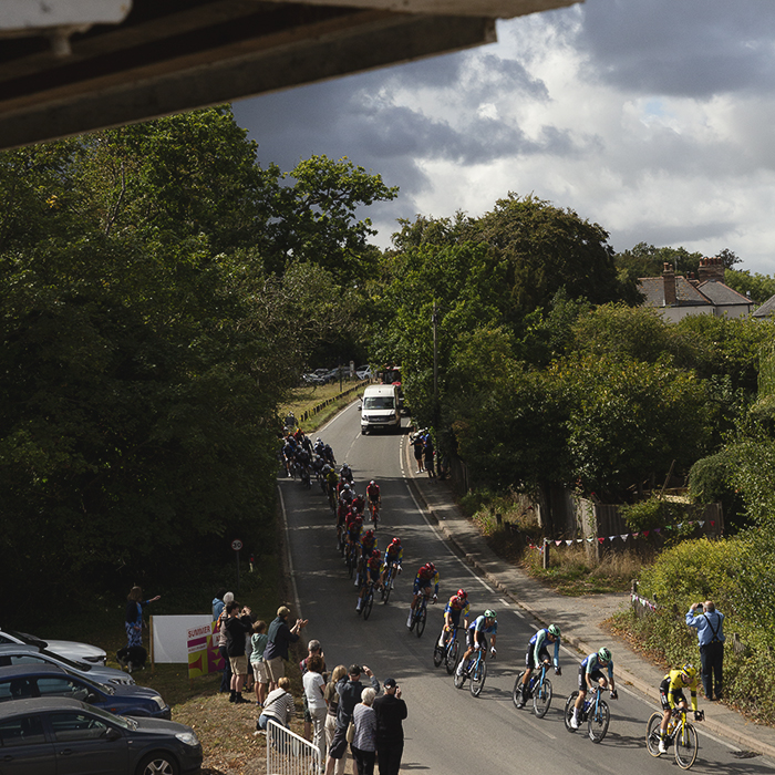 Tour of Britain 2025 - The peloton below the old warehouse buildings at Snape Maltings