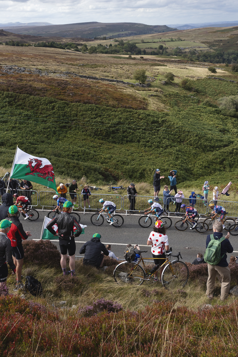 Tour of Britain 2025 - A fan sits on a bike wearing a polka dot jersey while others fly Welsh flags, a view of the hills as a backdrop