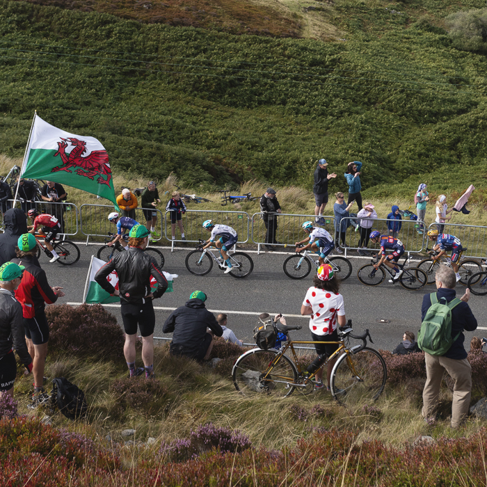 Tour of Britain 2025 - A fan sits on a bike wearing a polka dot jersey while others fly Welsh flags, a view of the hills as a backdrop