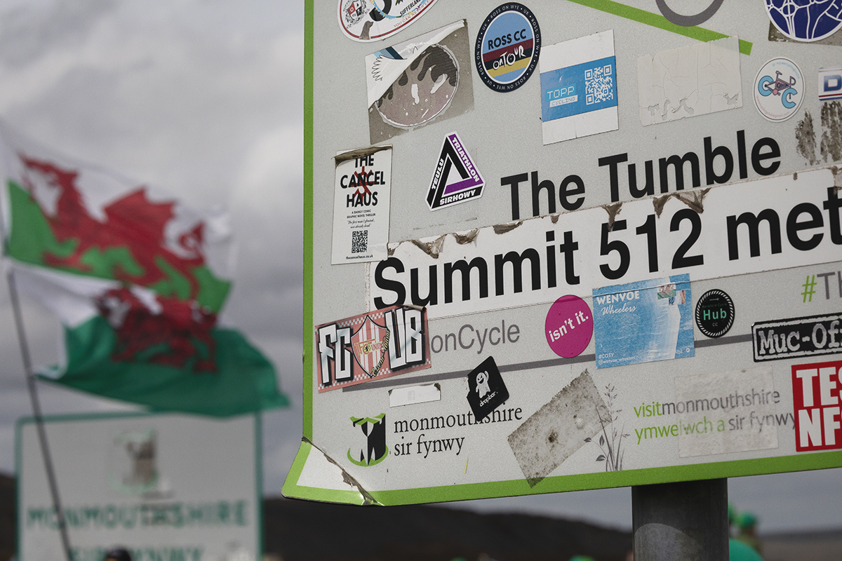 Tour of Britain 2025 - A sign for The Tumble covered in stickers, Welsh flags flying in the wind behind