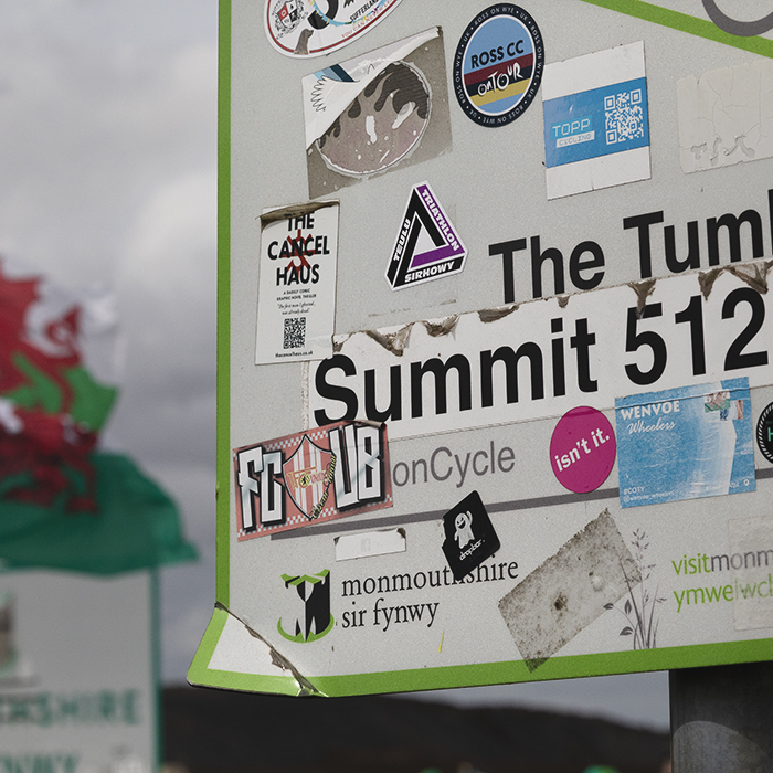 Tour of Britain 2025 - A sign for The Tumble covered in stickers, Welsh flags flying in the wind behind
