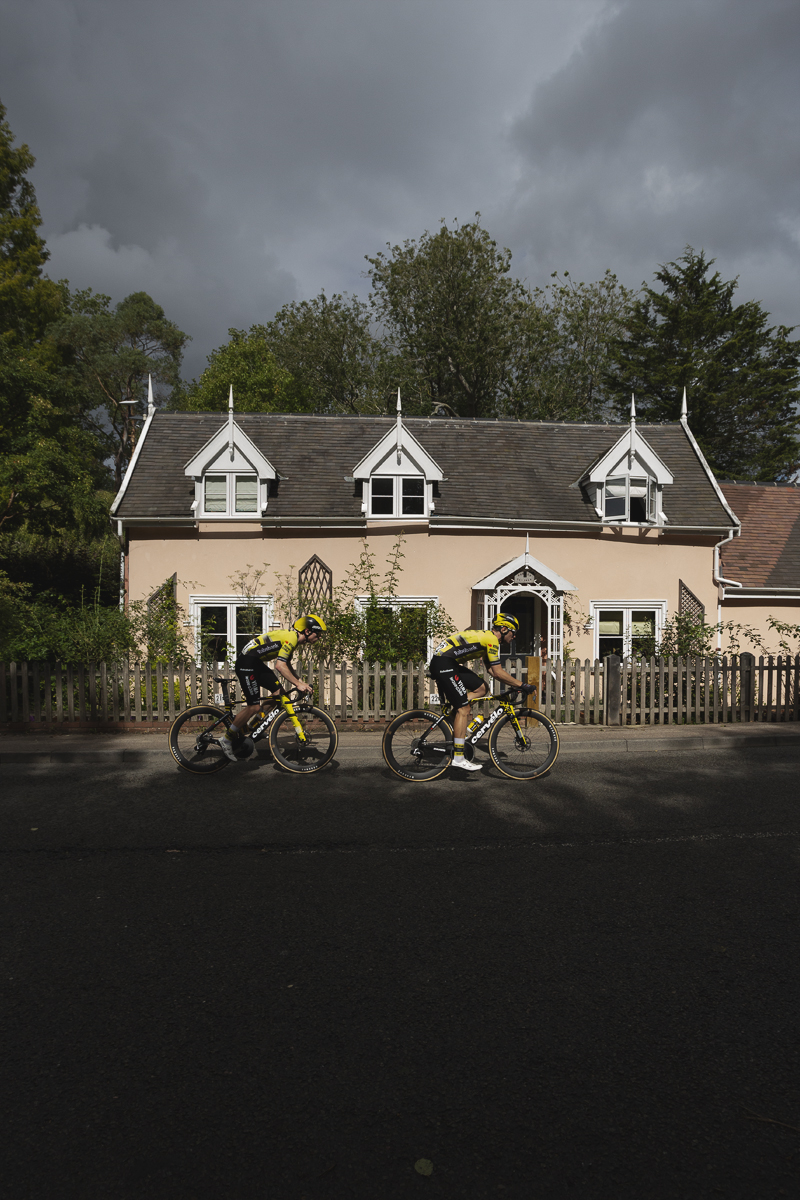 Tour of Britain 2025 - Matthew Brennan & Olav Kooij ride in front of a pastel coloured cottage in Walpole