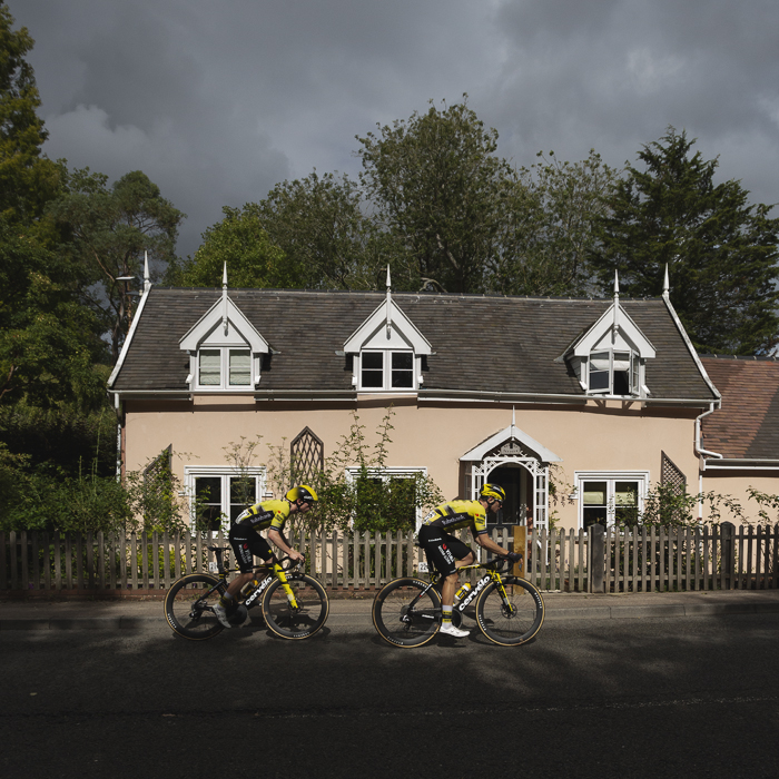 Tour of Britain 2025 - Matthew Brennan & Olav Kooij ride in front of a pastel coloured cottage in Walpole