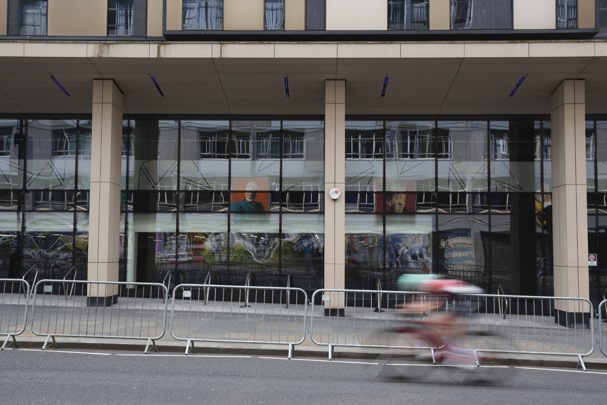 Tour of Britain Women’s 2025 - Stage 4 - A mural of the famous alumni of the University of Strathclyde is reflected in windows as a rider chases the race