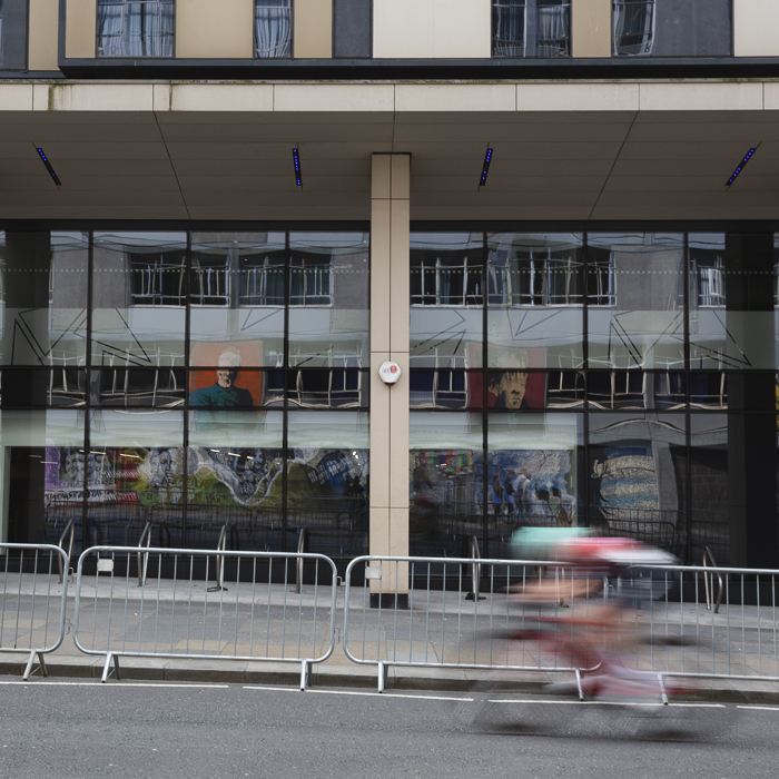 Tour of Britain Women’s 2025 - Stage 4 - A mural of the famous alumni of the University of Strathclyde is reflected in windows as a rider chases the race
