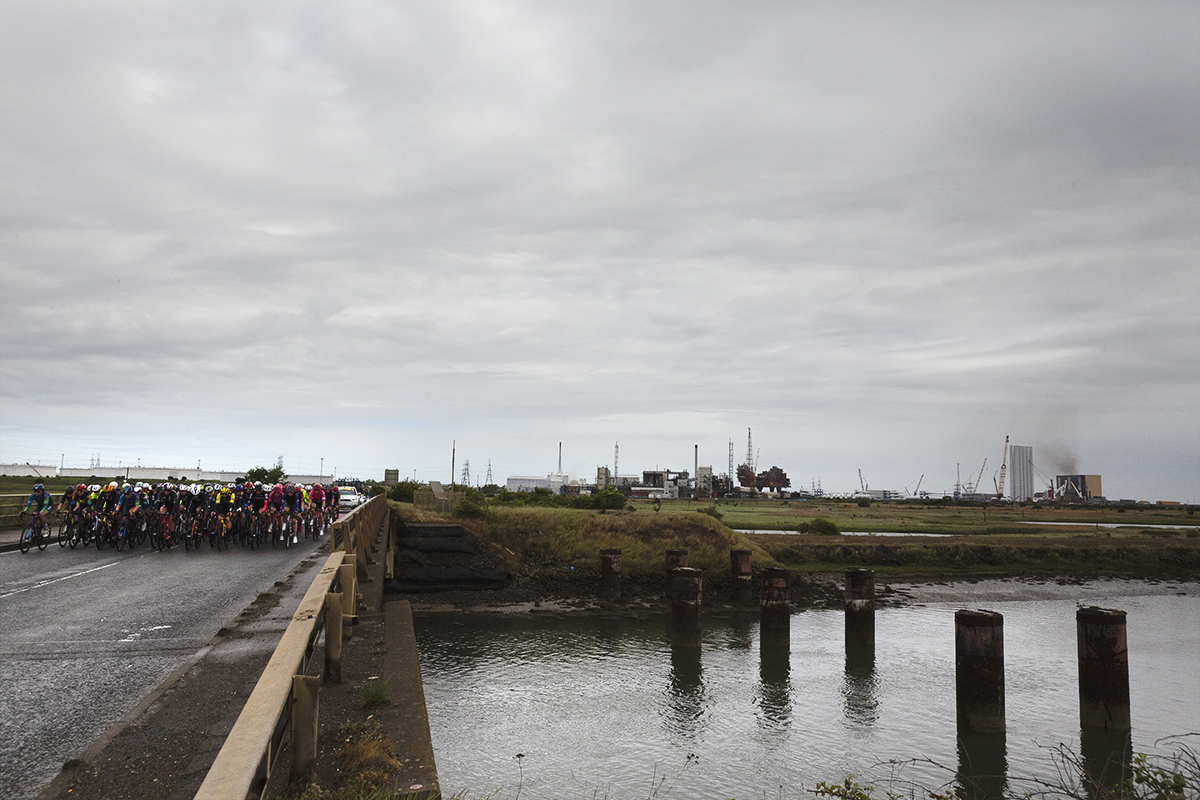 Tour of Britain Women’s 2025 - Stage 2 - The peloton on a bridge over Greatham Creek with industrial landscape in the background