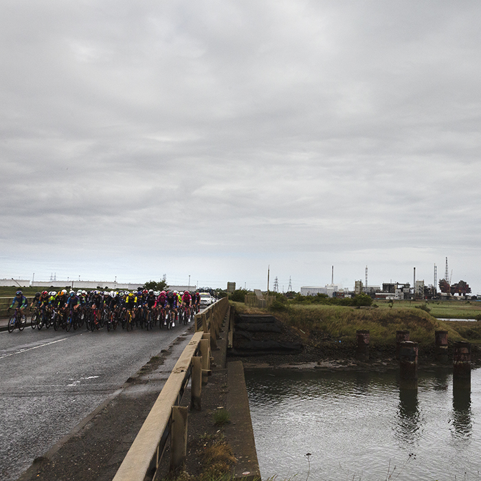 Tour of Britain Women’s 2025 - Stage 2 - The peloton on a bridge over Greatham Creek with industrial landscape in the background