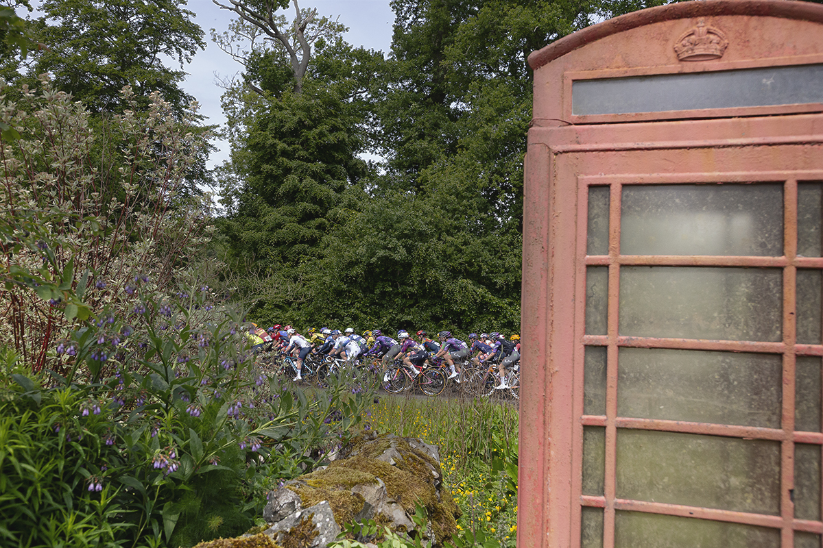 Tour of Britain Women’s 2025 - Stage 3 - The peloton is framed by a faded telephone box and a traditional cottage garden in Mellerstain