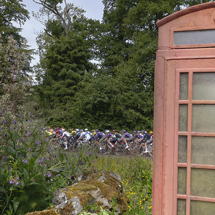 Tour of Britain Women’s 2025 - Stage 3 - The peloton is framed by a faded telephone box and a traditional cottage garden in Mellerstain