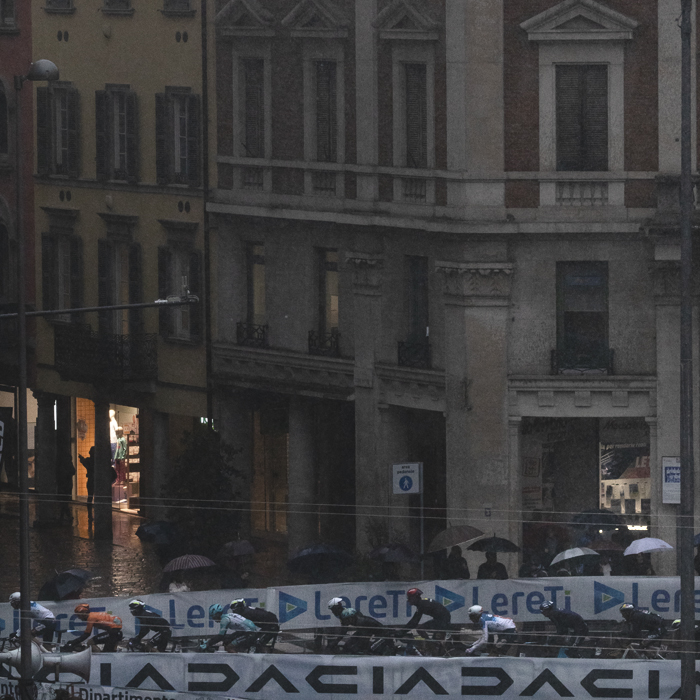 Tre Valli Varesine 2024 - A few hardy souls watch from under umbrellas and in shop doorways as the peloton passes majestic buildings in Varese.