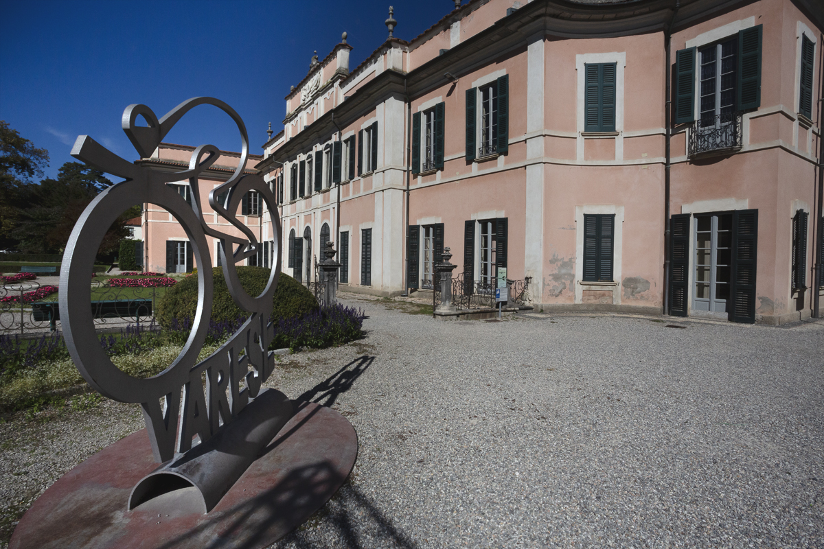 Tre Valli Varesine 2025 - A statute of a cyclist in a tuck position crafted from metal stands in the courtyard of a grand pink coloured building in Varese