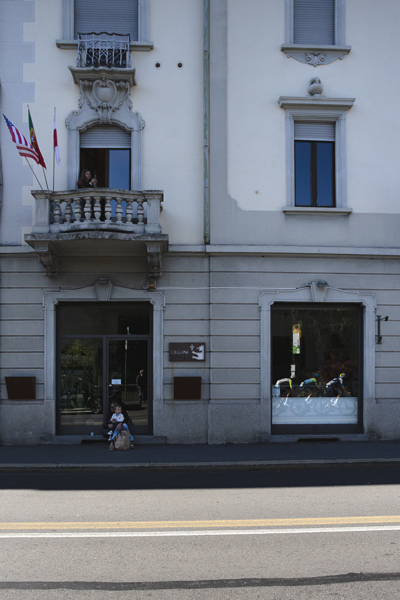 Tre Valli Varesine 2025 - A woman and child sit on the steps of a building to watch the race which is reflected in its windows