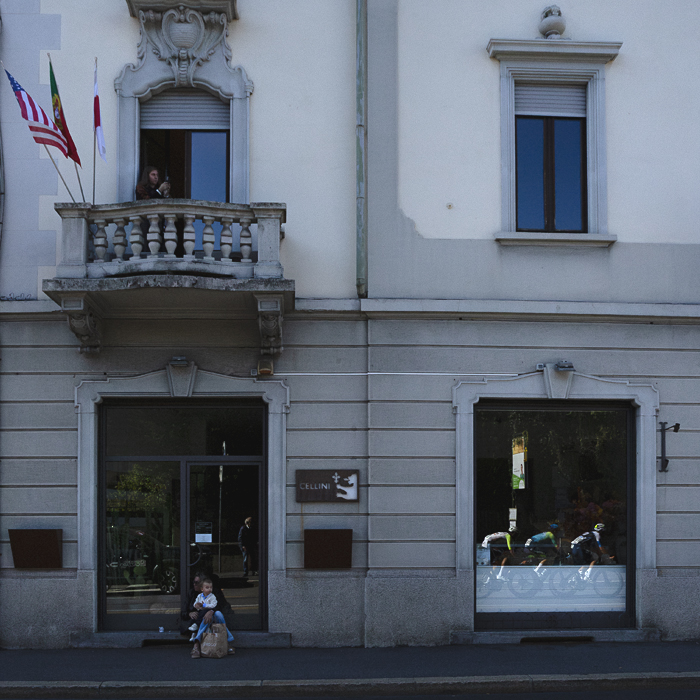 Tre Valli Varesine 2025 - A woman and child sit on the steps of a building to watch the race which is reflected in its windows