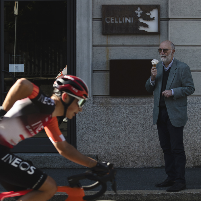 Tre Valli Varesine 2025 - A man with a beard eats a gelato while watching a rider pass by who is reaching for a gel
