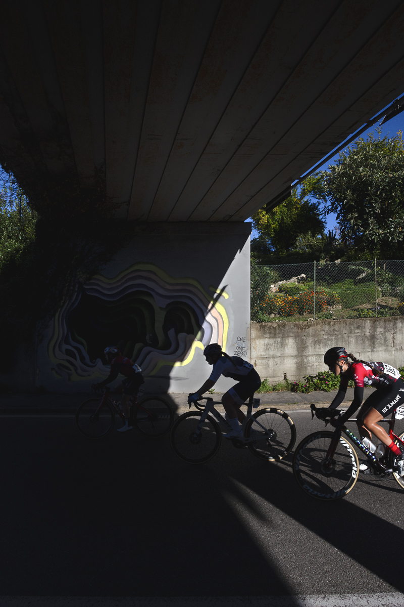 Tre Valli Varesine Women’s Race 2025 - Riders pass into the shadow of a modern road bridge which is decorated with an abstract mural