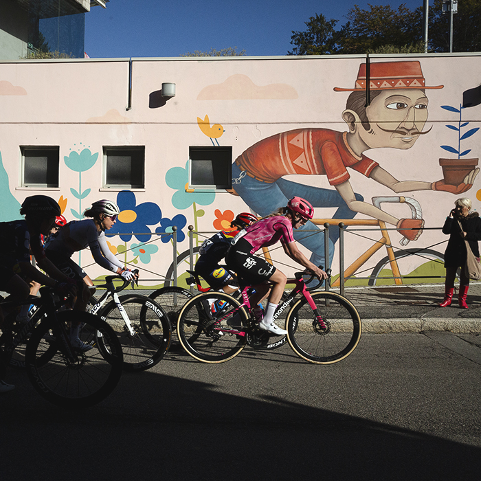 Tre Valli Varesine Women’s Race 2025 - Mirre Knaven of EF Education-Oatly leads a group of riders out of the shadows past a cheerful mural of a man in a hat holding a plant whilst riding a bike