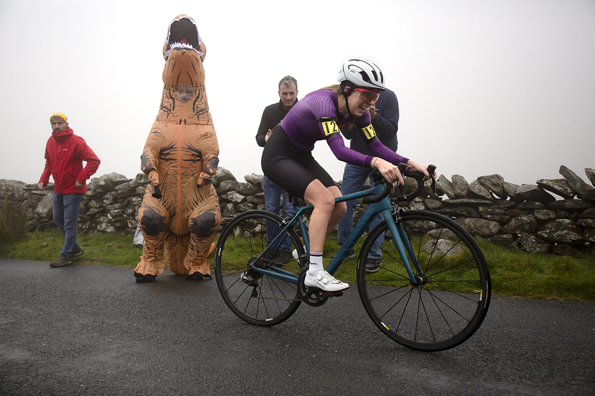UK National Hill Climb Championships 2023 - Amelia Cass passes a fan dressed as a dinosaur roaring at the sky as she makes her way up the climb