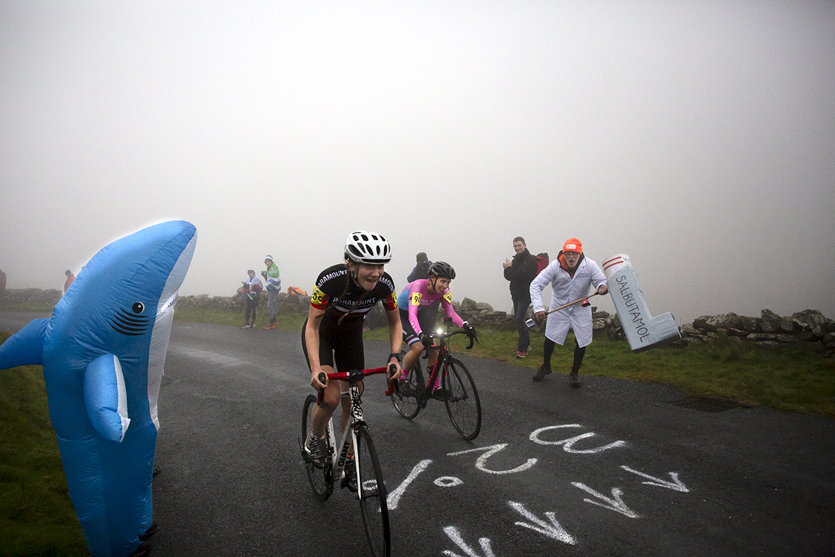 UK National Hill Climb Championships 2023 - Sara Willhoit & Rebecca Holland pass a fan with a giant inhaler in a lab coat and a fan dressed as a shark with 32% written on the road