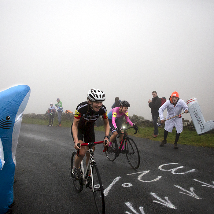 UK National Hill Climb Championships 2023 - Sara Willhoit & Rebecca Holland pass a fan with a giant inhaler in a lab coat and a fan dressed as a shark with 32% written on the road